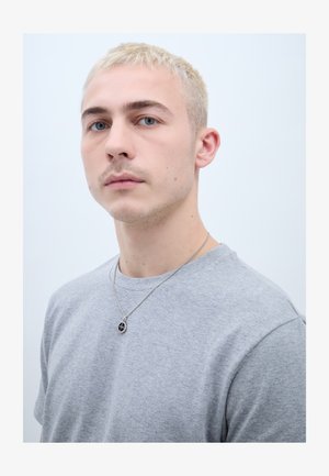 Noormees lühikeste heledate pleegitatud blondide juustega, kes kannab helehall T-särki ja hõbedast kaelakeed musta ripatsiga, valge ühevärvilise taustaga.