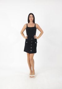 Kobieta stoi z rękami na biodrach, ubrana w czarny bezrękawnik, czarną spódnicę zapinaną na guziki z kieszeniami, białe sandały na obcasie z odkrytymi palcami oraz z tatuażami na nogach i ramieniu.