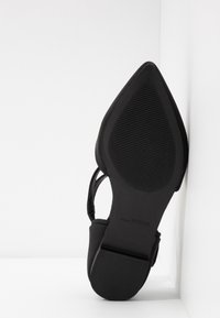 Zwarte hoge hakken met een puntige neus, gladde textuur en verstelbare band. Rubberen zool met ribbelstructuur voor grip.