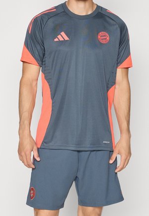 Homme portant un maillot de sport gris foncé et orange du FC Bayern Munich et un short assorti, debout avec les bras relâchés.