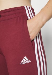 Tmavě červené sportovní kalhoty s bílým designem tří pruhů po stranách, s elastickým pasem a boční kapsou. Bílé logo Adidas.