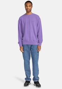 Felpa a girocollo viola con collare e polsini a coste, abbinata a jeans in denim azzurro chiaro e sneakers nere.