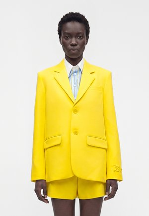 Personne portant un blazer jaune vif et un short assorti sur une chemise à rayures bleues et blanches, se tenant devant un fond uni.