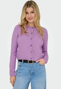 Lila stickad kofta med långa ärmar, rund halsringning och svarta knappar framtill. Kombinerad med högmidjade blåjeans.