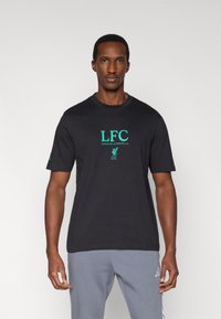 Czarny bawełniany t-shirt z zielonym logo "LFC", krótkimi rękawami i okrągłym dekoltem. Noszony z szarymi spodniami dresowymi.