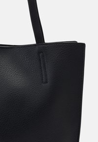 Černá kožená taška typu tote s texturovaným povrchem, které má strukturovaný tvar a dvě pevné rukojeti. Minimalistické prošívání na vrchu.