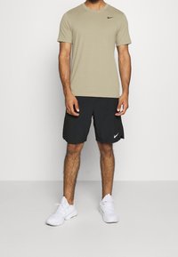 Camisa de manga corta verde oliva para hombre con un logo negro de Nike, combinada con pantalones cortos negros que tienen un logo blanco de Nike y zapatillas blancas.