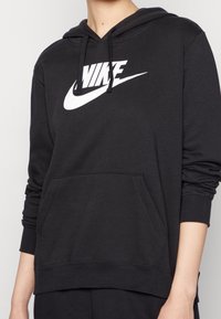 Person som bär en svart Nike-hoodie med vitt logo, framficka och snoddar, torso och armar synliga mot en neutral bakgrund.