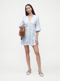 Femme portant une robe bleu clair à motifs avec des manches bouffantes, tenant un sac à main marron et portant des chaussures blanches à petit talon.