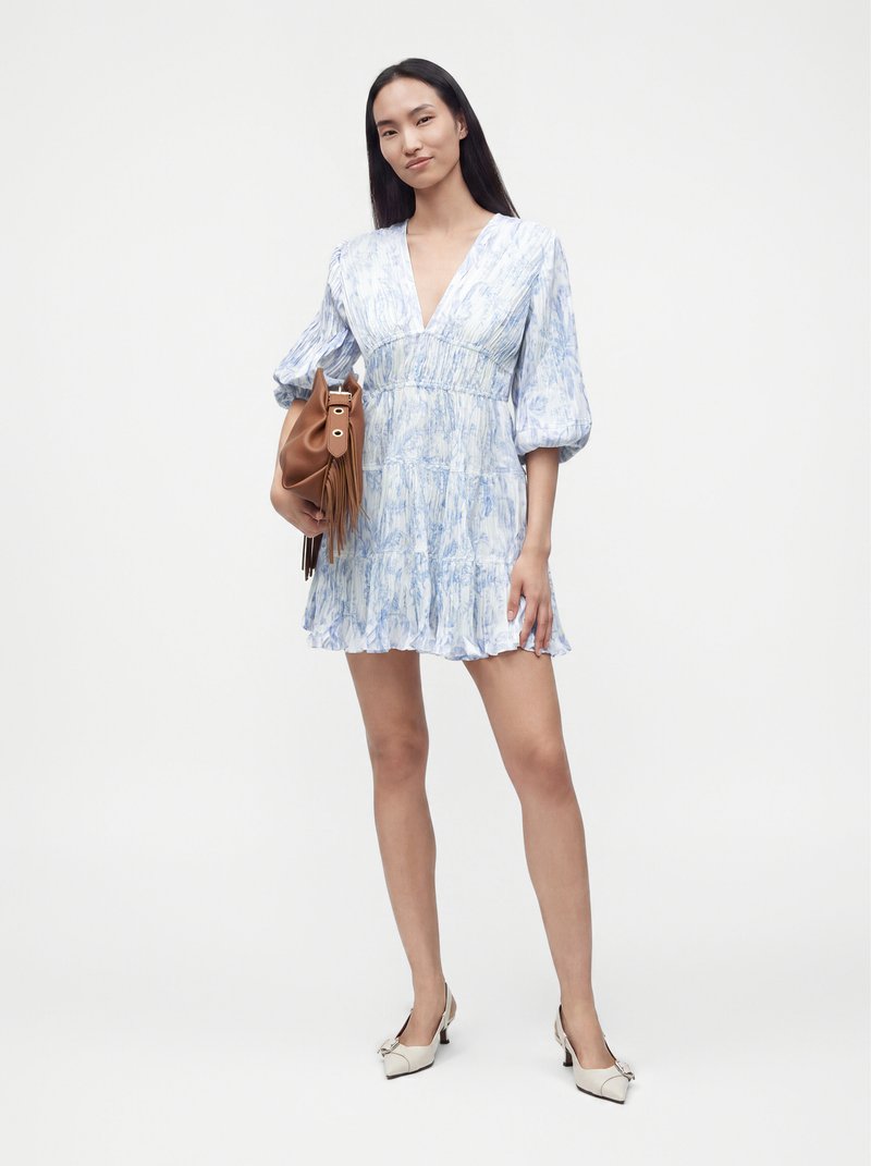 Woman wearing a light blue patterned dress with puffed sleeves, holding a brown handbag, and wearing white low-heeled shoes.