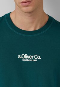 Türkisfarbiges Baumwoll-T-Shirt mit Rundhalsausschnitt und kurzen Ärmeln. Auf der Vorderseite befindet sich ein weißer Druck-Logo "s.Oliver Co. Established 1969".