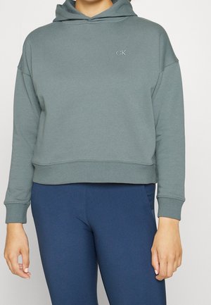 Vrouw draagt een cropped, gedempte groene hoodie met "CK"-logo op de borst en nauwsluitende marineblauwe broek, staand tegen een effen achtergrond.