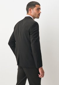 Schwarze maßgeschneiderte Anzugjacke mit schmaler Passform, mit Reverskragen, einem Schlitz hinten und geknöpften Manschetten, getragen über einer passenden Hose.