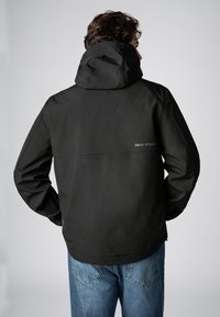 Zwarte hoodie met een gladde textuur, voorzien van een subtiel logo op de achterkant, gecombineerd met losse blauwe denim jeans.