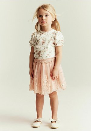 Blondes Mädchen trägt ein geblümtes Oberteil, einen rosa Spitzenrock und weiße Schuhe mit Schleifen, steht vor einem schlichten hellen Hintergrund.