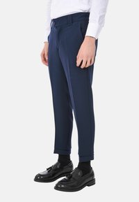 Pantaloni navy sartoriali con frontale piatto, tasche laterali e piega accentuata. Indossati con scarpe e calzini neri, dotati di chiusura a bottoni.