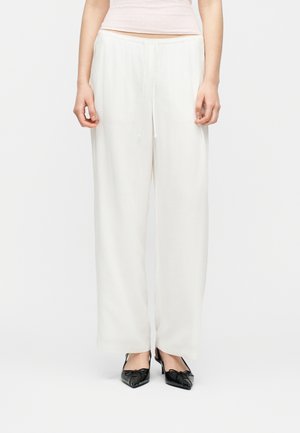 Žena v bielych nohaviciach s vysokým pásom a širokými nohavicami, so šnúrkou v páse a čiernymi lodičkami s ostrou špičkou, stojaca na jednoduchom pozadí.