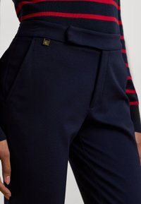 Navy trousers made of smooth fabric, featuring side pockets, a belted waist, and a small gold logo detail on the waistband.