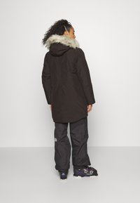 Parka marrón con capucha de piel sintética, diseño de largo medio y acabado mate. Combinada con pantalones de exterior negros y botas de senderismo negras.