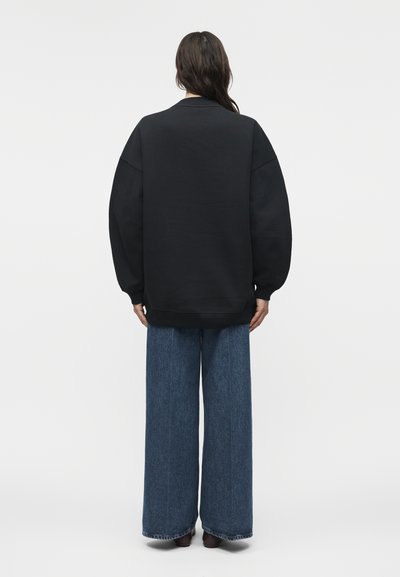 Άτομο με μακριά σκούρα μαλλιά στέκεται με την πλάτη, φορώντας μία φαρδιά μαύρη φούτερ μπλούζα και μπλε τζιν παντελόνι με φαρδιά πόδια, μπροστά σε ένα απλό φόντο.