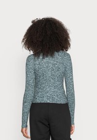 Blusa de manga longa em tecido de malha texturizado azul e preto, de design ajustado com uma bainha arredondada, vestida com calças pretas, mostrando a vista traseira.