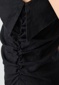 Abito in lino nero con colletto a punta e dettagli arricciati lungo il lato. Il tessuto appare testurizzato e leggermente trasparente.