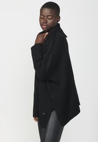 Črn oversized pulover z teksturiranim pletenjem, z visokim ovratnikom, stranskimi razpoki in rebrastimi manšetami. V kombinaciji z elegantnimi črnimi legicami.