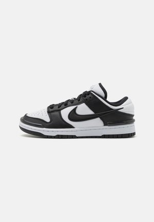 Lavtopet sneaker i sort og hvidt læder med sort Nike swoosh og sorte snørebånd på hvid sål mod ensfarvet grå baggrund.