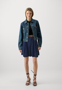 Jachetă din denim cu nasturi și buzunare în față, top scurt negru, fustă fluidă de culoare bleumarin și sandale negre cu curele. Texturi fine și siluetă casual.