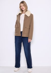 Giacca in misto lana marrone con colletto in pelliccia sintetica, due tasche frontali, abbinata a jeans blu e sneaker bianche, in piedi su uno sfondo bianco.