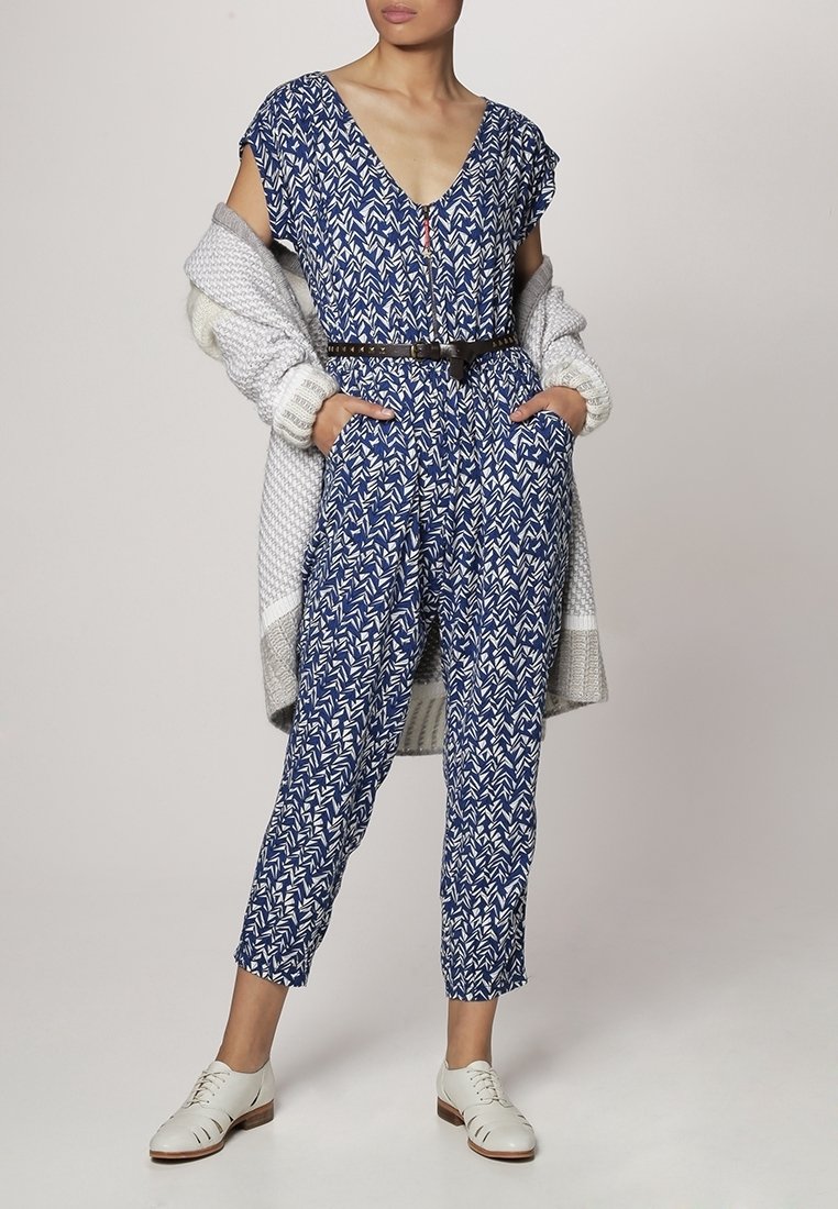 Femme portant une combinaison bleue et blanche à motifs, une ceinture noire, un cardigan en tricot gris sur les épaules, et des chaussures blanches à lacets.
