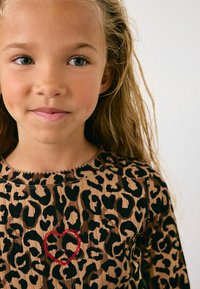 Noor tüdruk, kellel on laineline kuldne juuksed, seljas pruun leopardimustriga topp, millel on rinnal väike punane tikandiga süda, ühtlases taustas.