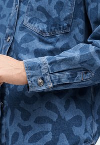 Jeanshemdärmel mit dunkelblauen abstrakten Mustern, Metallknopfmanschette und sichtbaren Nähten der Brusttasche am Arm einer Person.