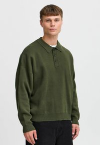 Mørkegrønn strikket polo med krage, tre knapper og ribbede mansjetter, kombinert med mørke jeans. Subtil tekstur gjennom hele plaggene.