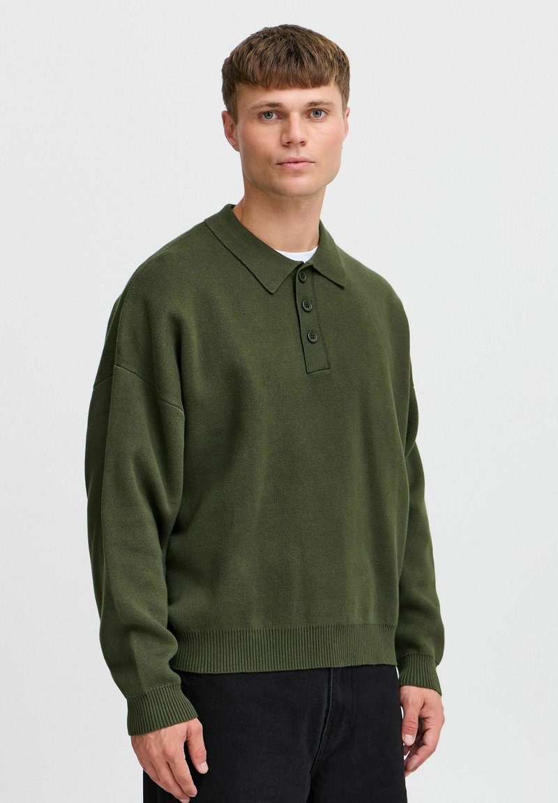 Mørkegrønn strikket polo med krage, tre knapper og ribbede mansjetter, kombinert med mørke jeans. Subtil tekstur gjennom hele plaggene.