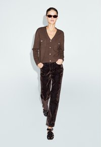 Cardigan marrone con bottoni frontali e due tasche sul petto, abbinato a pantaloni di velluto neri e sandali neri con cinturini, su uno sfondo chiaro.
