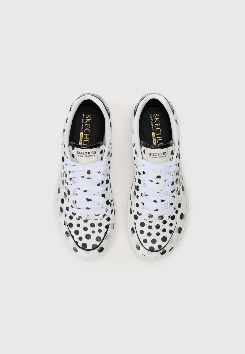 Une paire de baskets blanches Skechers à lacets avec des pois noirs et des semelles en mousse à mémoire, vue de dessus sur un fond uni.