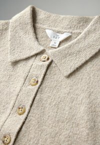 Bežová pletená polo košeľa s textúrou, vybavená golierom a štyrmi gombíkmi. Nálepka znie "THE SET", veľkosť M. Materiál pôsobí mäkko a teplo.
