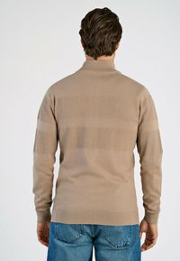Beżowy sweter dzianinowy z teksturowanym wzorem w paski, z wysokim kołnierzem i dopasowanym krojem. Noszony z niebieskimi dżinsami.