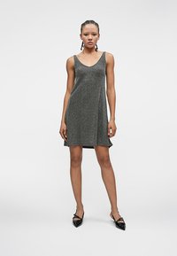 Fiatal nő, fonott hajjal, ujjatlan, csillogó sötétszürke ruhában és fekete, hegyes orrú lapossarkú cipőben áll, sima háttér előtt.