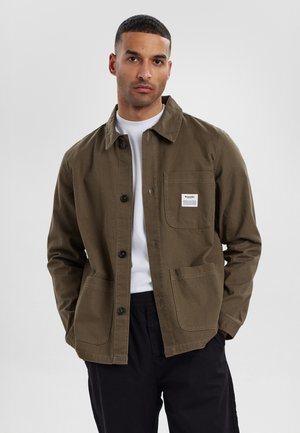 Brown cotton jacket with a collared neckline, front buttons, two large pockets, and a logo patch on the chest. Worn over a white shirt.