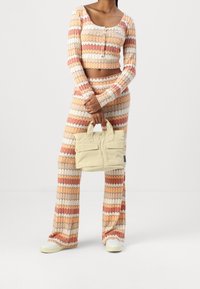Strikket to-delt sæt i orange, creme og røde vandrette striber; langærmet crop top med knapper, vide bukser; beige håndtaske med to lommer.
