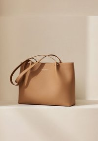 Sac fourre-tout en cuir tanné avec deux anses, une texture lisse et un design simple. Présentant des accessoires en métal doré et un logo subtil.