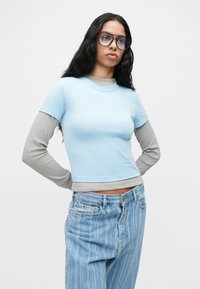 Νεαρή γυναίκα με μακριά σκούρα μαλλιά, που φοράει μπλε τόνων oversized γυαλιά, ανοιχτού μπλε κοντομάνικο μπλουζάκι πάνω από γκρι μακριά μανίκια και φαρδύ ριγέ τζιν.