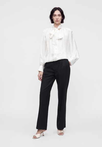 Femme debout portant un chemisier blanc à manches longues avec un nœud, un pantalon noir ajusté et des talons ouverts blancs sur un fond uni.
