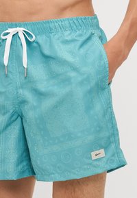 Törökzöld fürdőnadrág bandana mintával, rugalmas derékkal, húzózsinórral és oldalsó zsebekkel. Az anyag sima és könnyűnek tűnik.