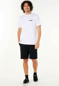 Weißes Baumwoll-T-Shirt mit dem 'RIP CURL'-Logo auf der Brust, kombiniert mit schwarzen Shorts. Modell trägt schwarze Sneakers und Knöchelsocken.