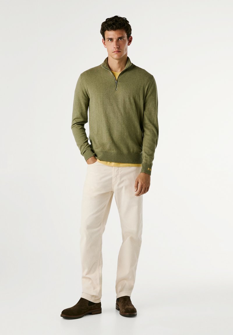 Zielony sweter zapinany na zamek na żółtej koszulce, beżowe spodnie i brązowe zamszowe botki. Stylizacja prezentuje swobodny, warstwowy wygląd z luźnym krojem.