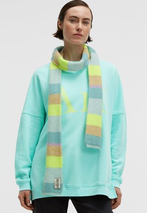 Frau mit einem pastellgestreiften Schal über einem hellblauen Sweatshirt mit gelber Schrift, stehend vor einem schlichten hellen Hintergrund.