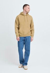 Beige hættetrøje med en forlommer, parret med blå jeans og beige sneakers. Stofmaterialet virker blødt med en afslappet pasform.